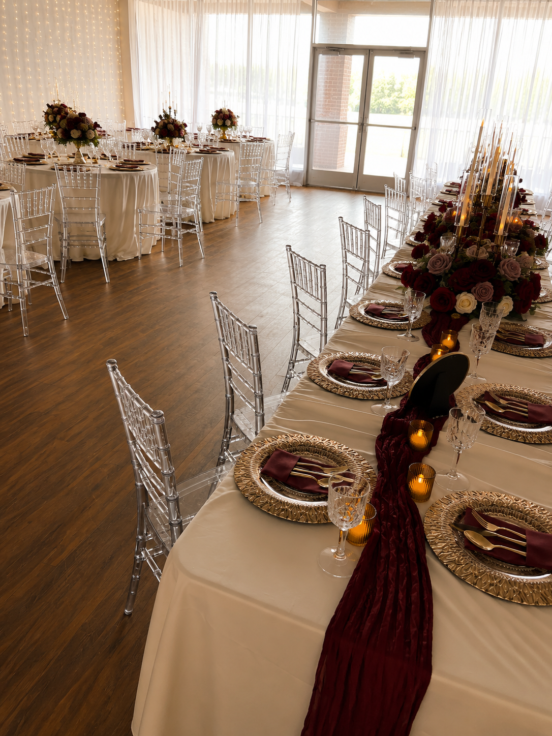 A long banquet table set in a banquet hall with white tablecloths, silver Chiari chairs, gold charger plates, burgundy napkins, and deep burgundy floral centerpieces. The room has a dark wood floor and large windows with sheer white curtains.